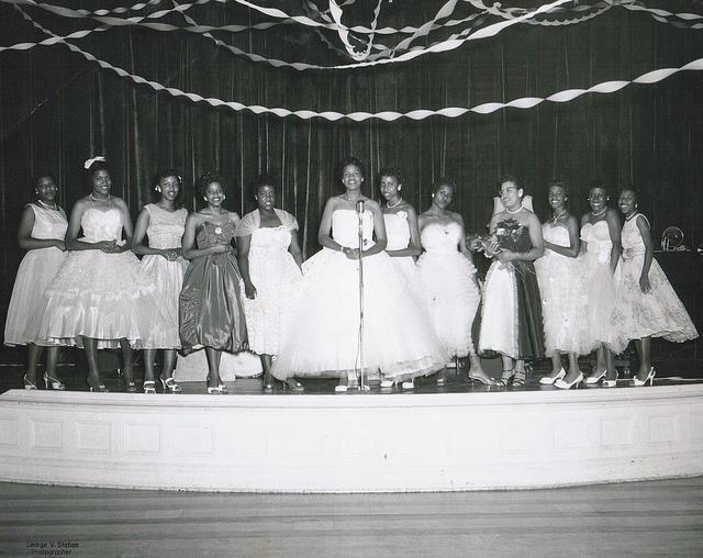 Young women in fancy dresses stand on a stage while one addresses the audience